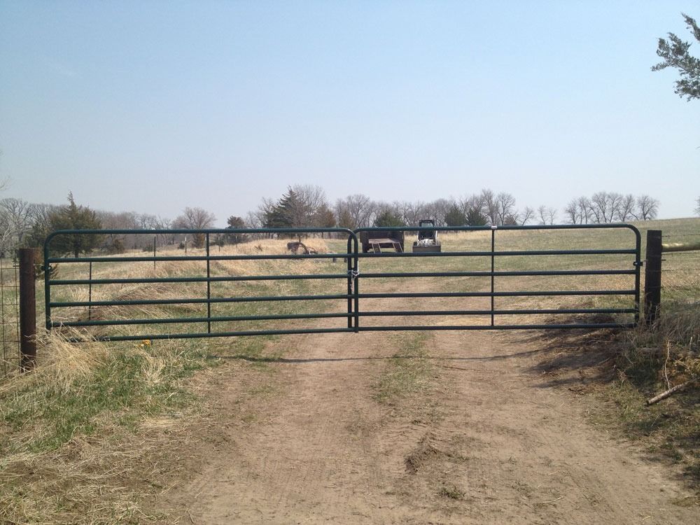 Green metal gate across a dirt road in a field; trees in background; sunny day.
