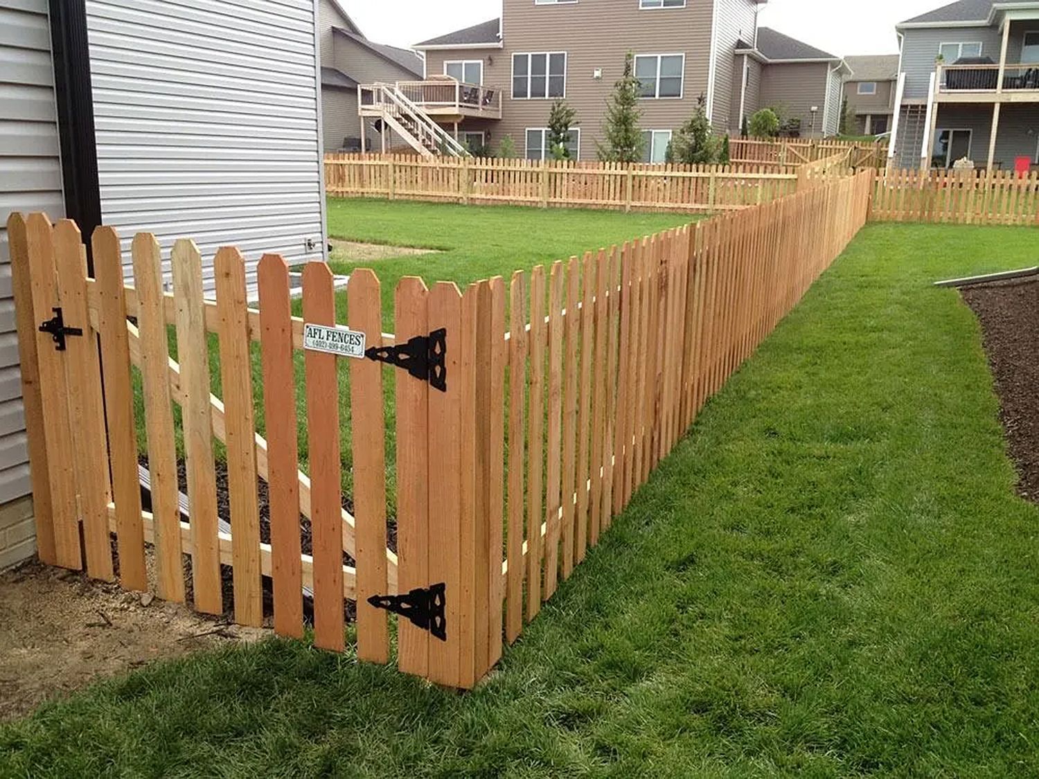 Wooden picket fence and gate surround a green yard with houses in the background.