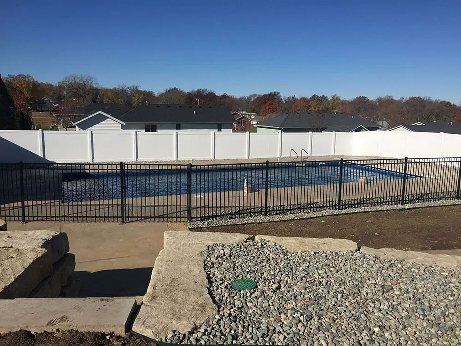 Pool with black metal fence, white privacy fence, and houses in the background under a blue sky.