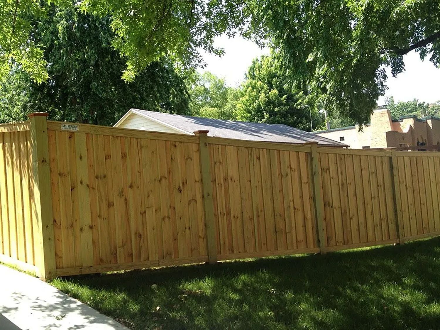 Wooden fence in a yard, with green grass in the foreground and trees and a roof in the background.