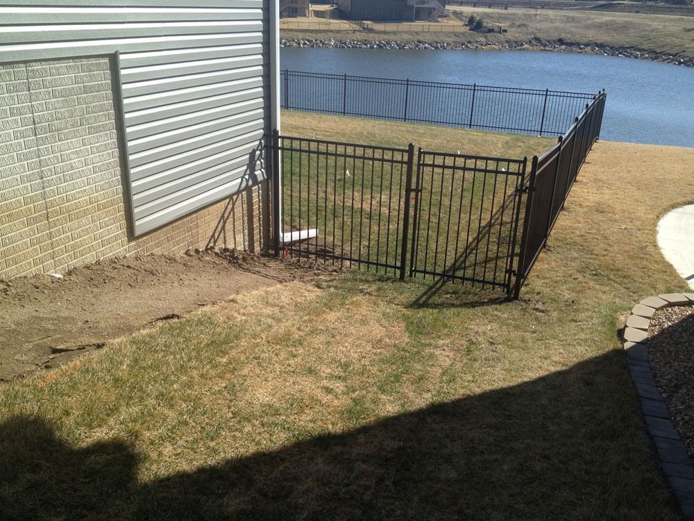 Black metal fence section near a building and a lake. Brown grass and dirt are nearby.