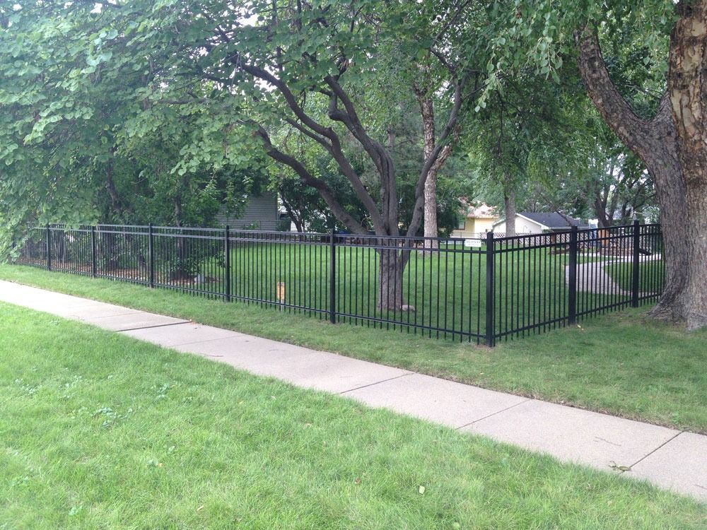 Black metal fence around a grassy yard with trees, next to a sidewalk.