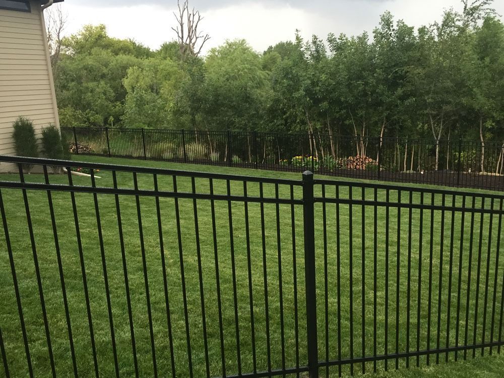Black metal fence encloses a green lawn with a wooded area in the background under a cloudy sky.