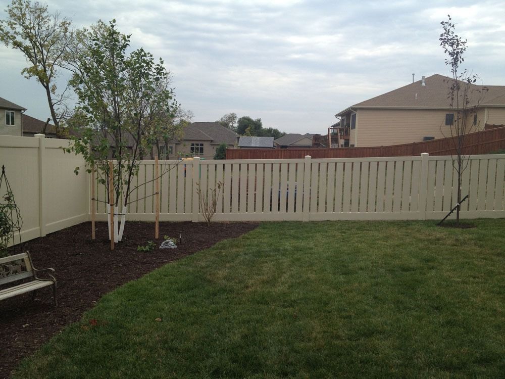Backyard with green grass, brown mulch, beige fence, and small trees under a cloudy sky.