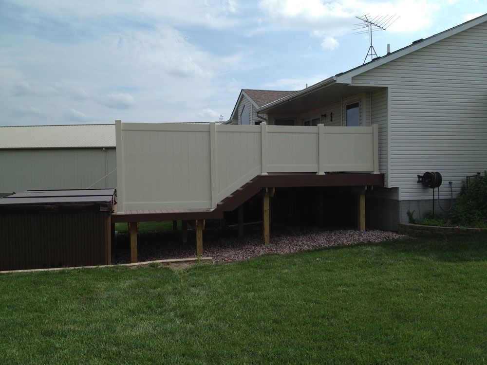 Brown deck with stairs, tan fence, and hot tub. House in the background. Green grass and blue sky.