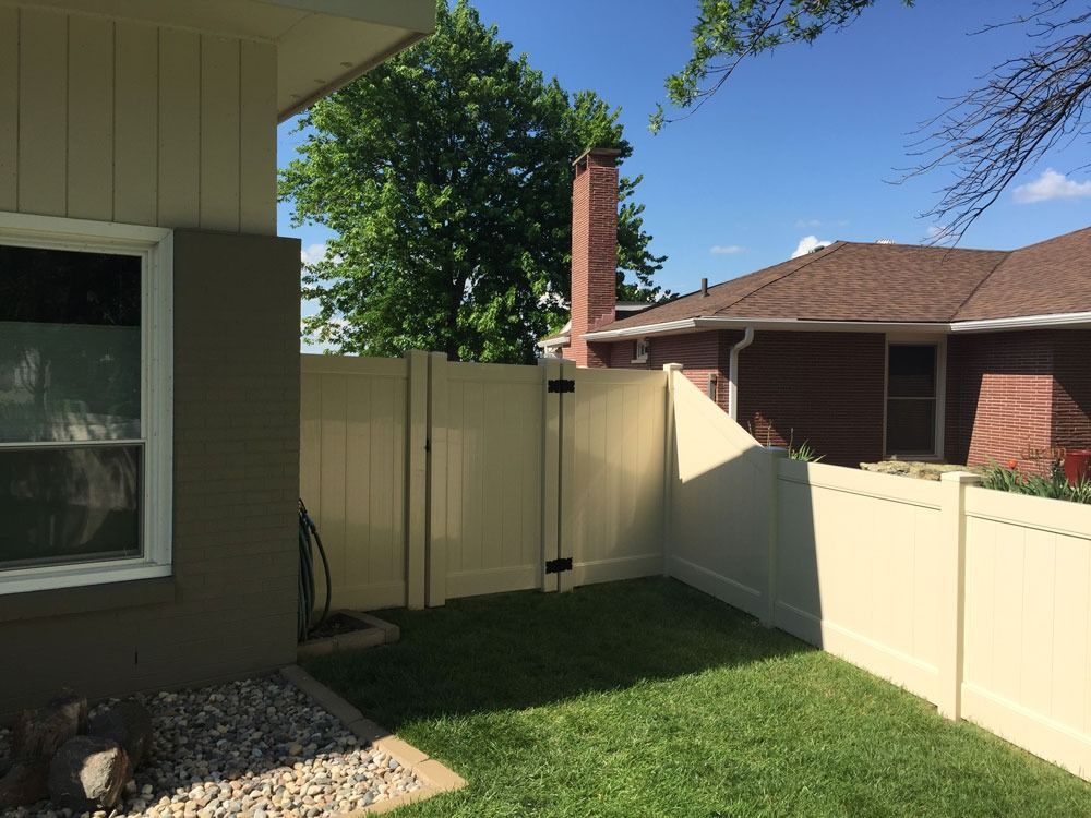 Beige vinyl fence enclosing a small grassy yard beside a house with a window. Sunny day.