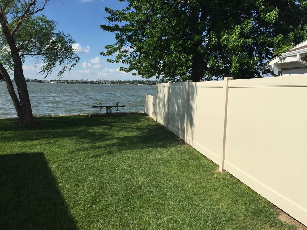 Lawn with a fence by water. A beige fence borders a grassy yard, with a lake and trees in the background.