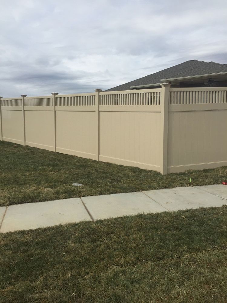 Beige vinyl fence enclosing a grassy yard, sidewalk in foreground, overcast sky.
