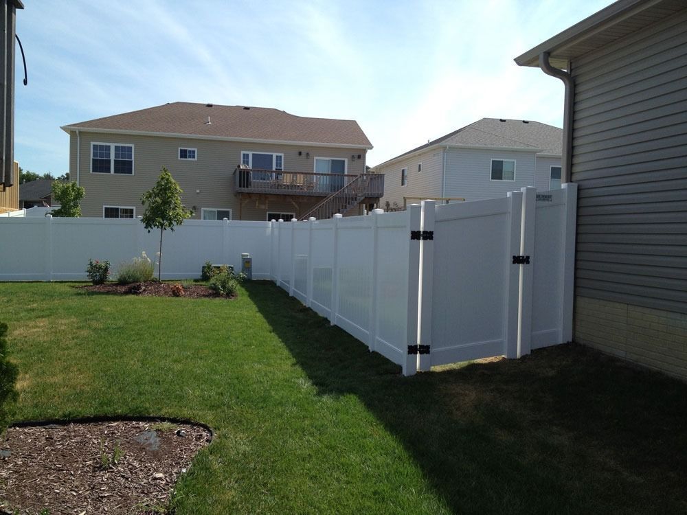 White vinyl fence and gate in a backyard, separating a grassy lawn from neighboring houses on a sunny day.