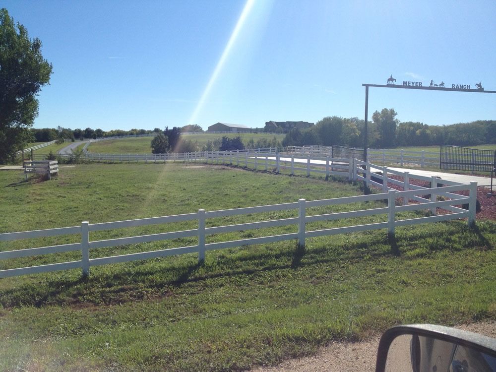 White fence lines a grassy field leading to an arched gate under a bright blue sky.