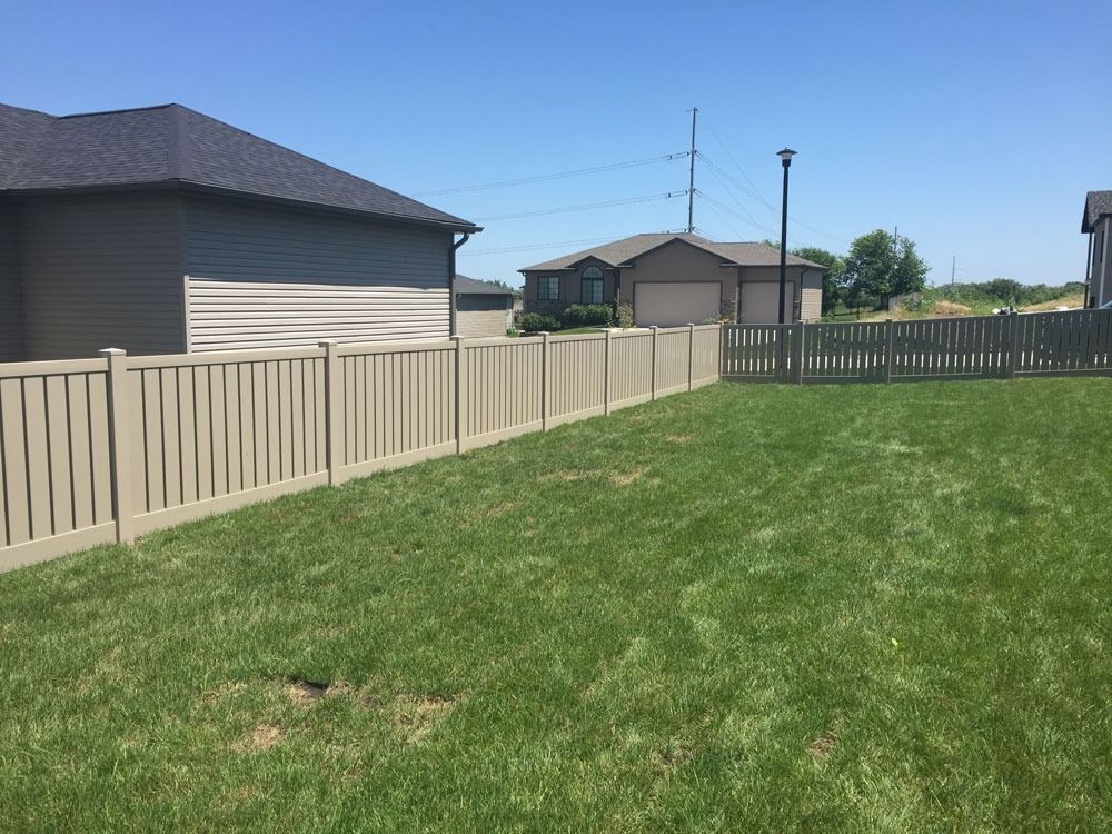 Tan vinyl fence encloses a green lawn in a suburban backyard under a blue sky.