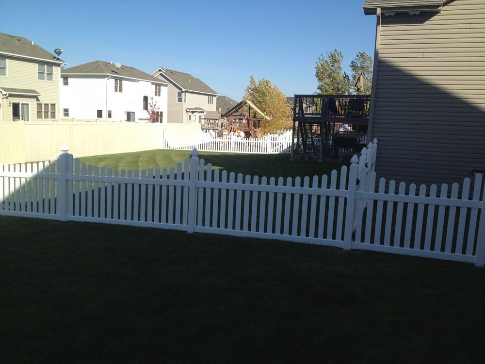 A white picket fence encloses a green backyard, with houses in the background under a blue sky.