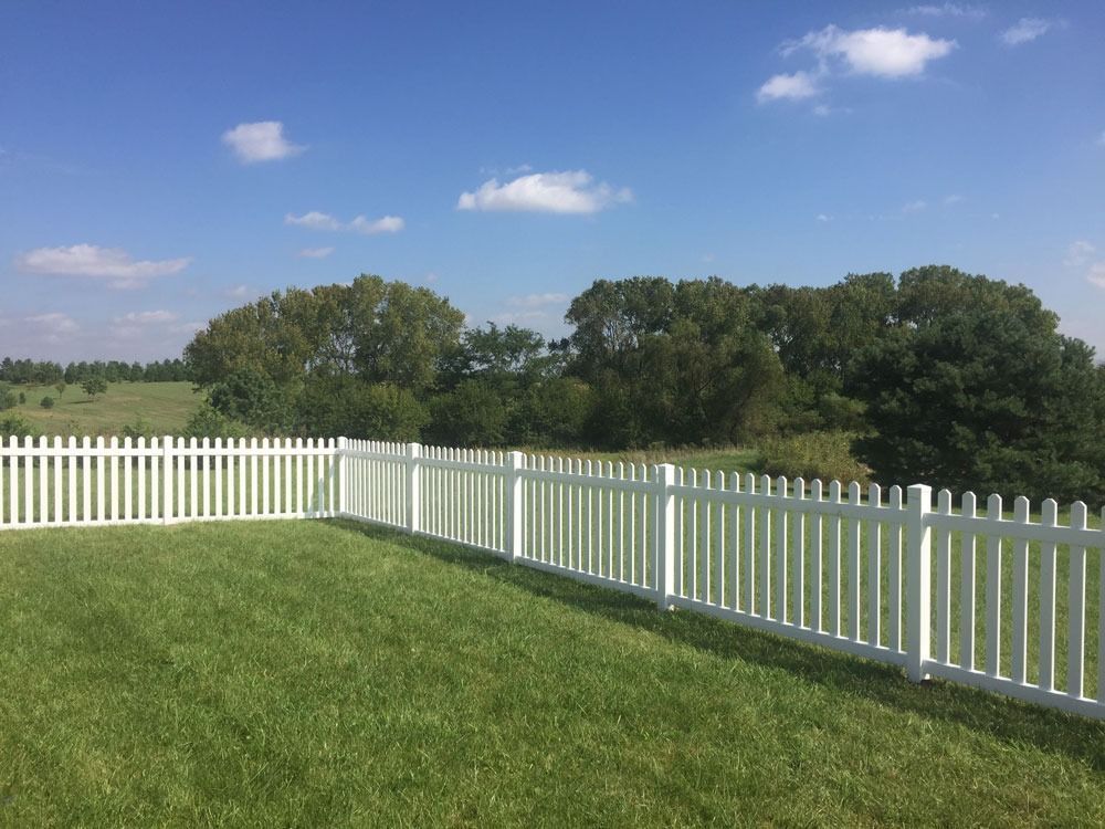 A white picket fence surrounds a green lawn against a backdrop of trees, hills, and blue sky with clouds.