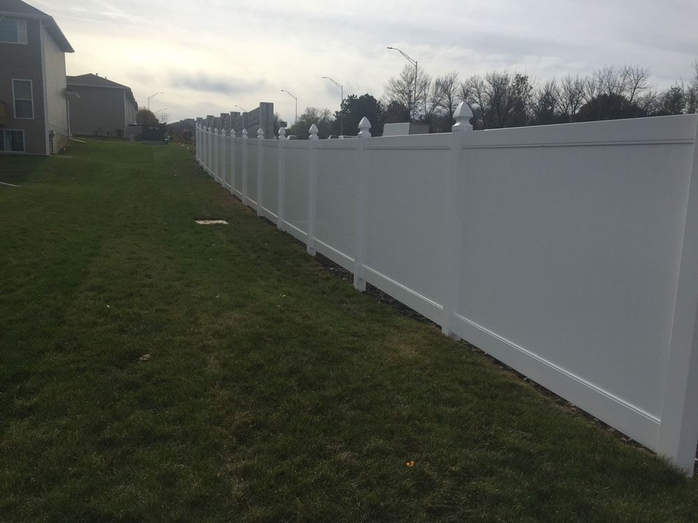 White vinyl fence along grassy yard, houses in the background under an overcast sky.