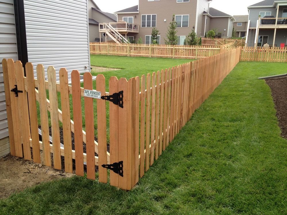 Wooden picket fence and gate surrounding a green yard, adjacent to a house with beige siding.
