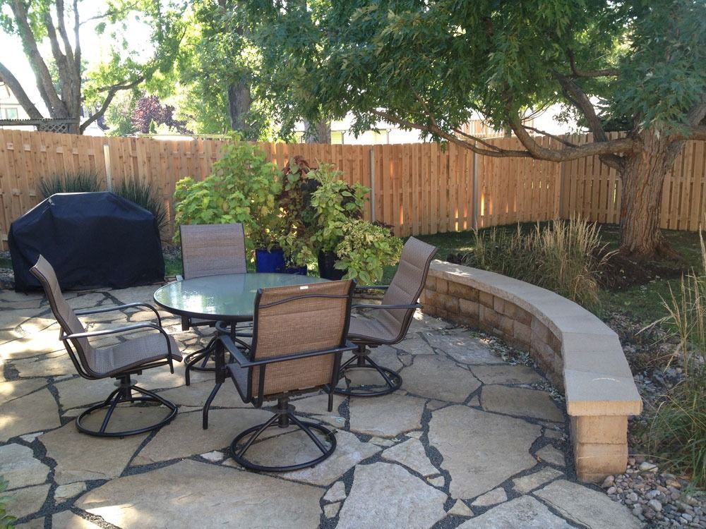Patio with a round table, swivel chairs, a curved stone bench, a wooden fence, and trees.