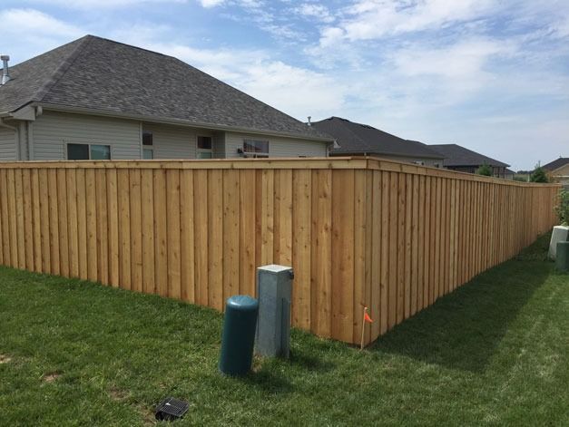A wooden privacy fence surrounds a grassy backyard, adjacent to a residential building under a partly cloudy sky.