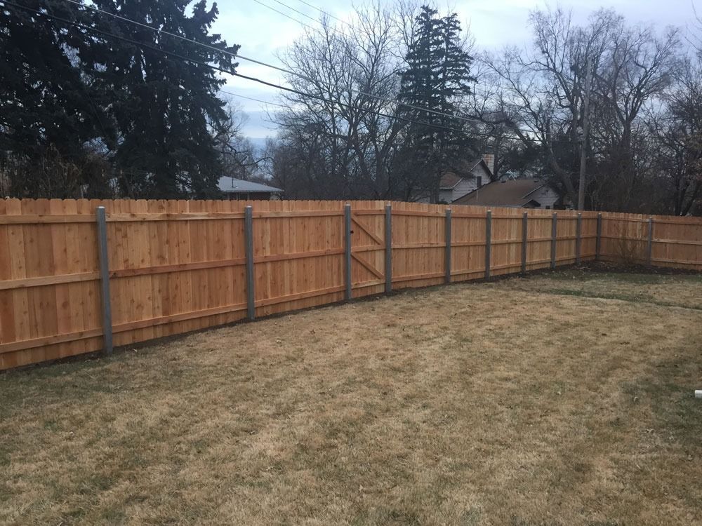 Wooden fence surrounds a yard with dry grass, trees, and a cloudy sky in the background.