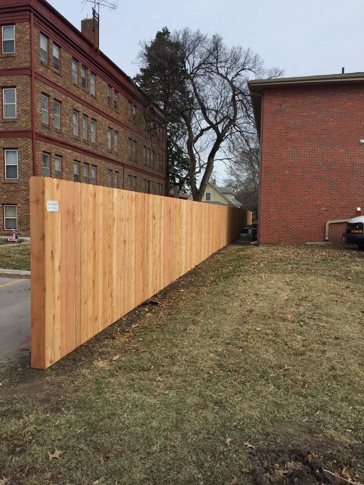 Tall wooden fence between brick buildings, on a grassy patch.