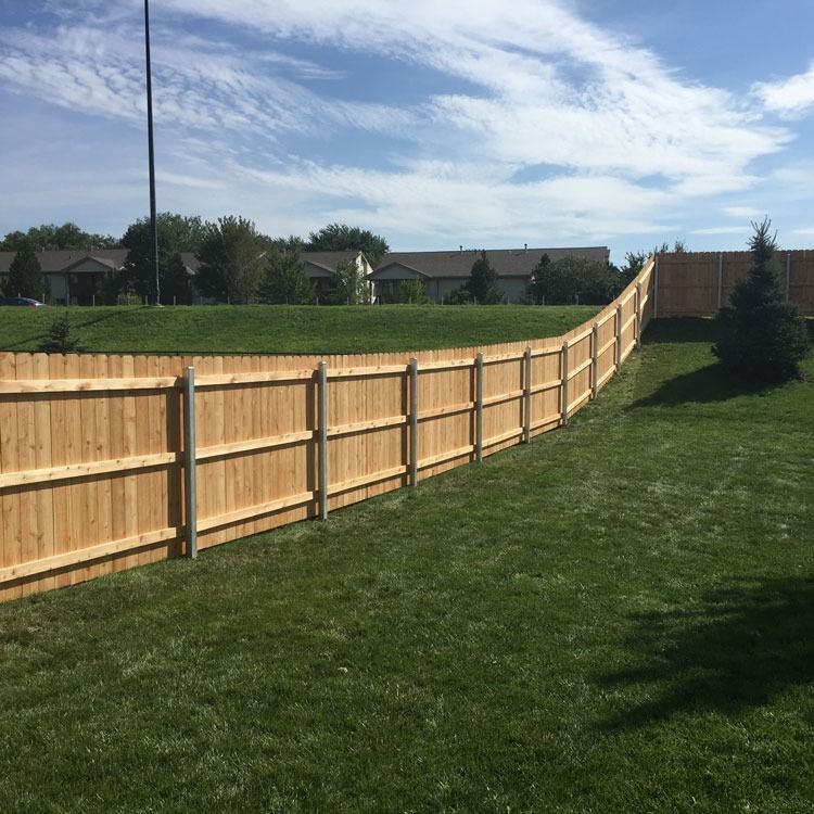 Wooden fence curving across a grassy yard under a blue sky, with houses in the background.