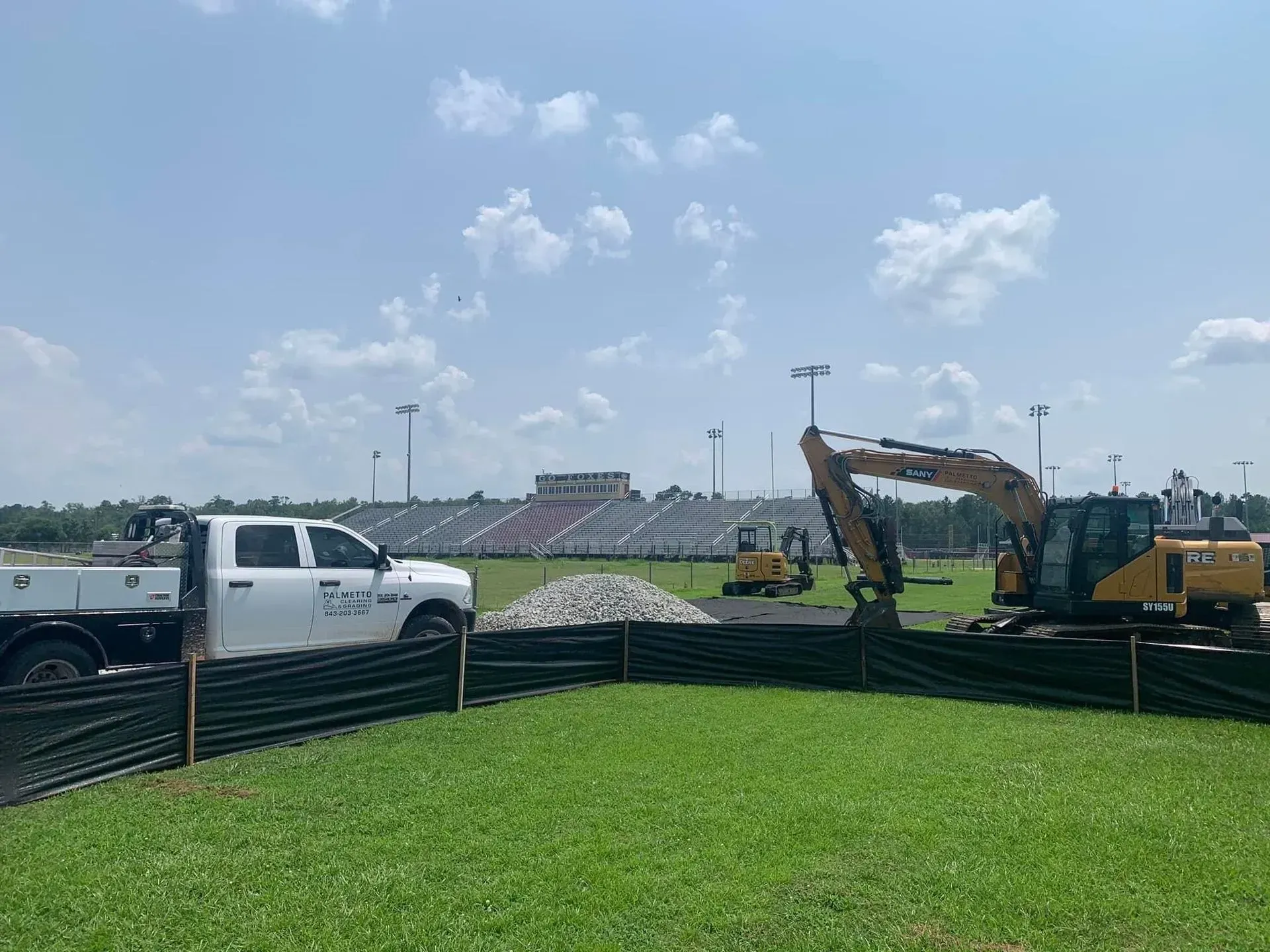 Construction site: white truck, excavator, and stadium in background. Green grass and blue sky.