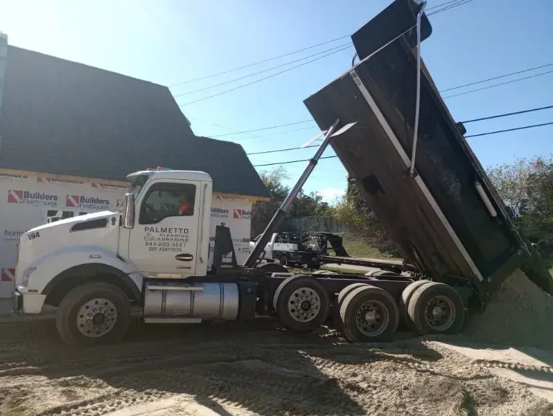 White dump truck dumping gravel next to a house under construction on a sunny day.