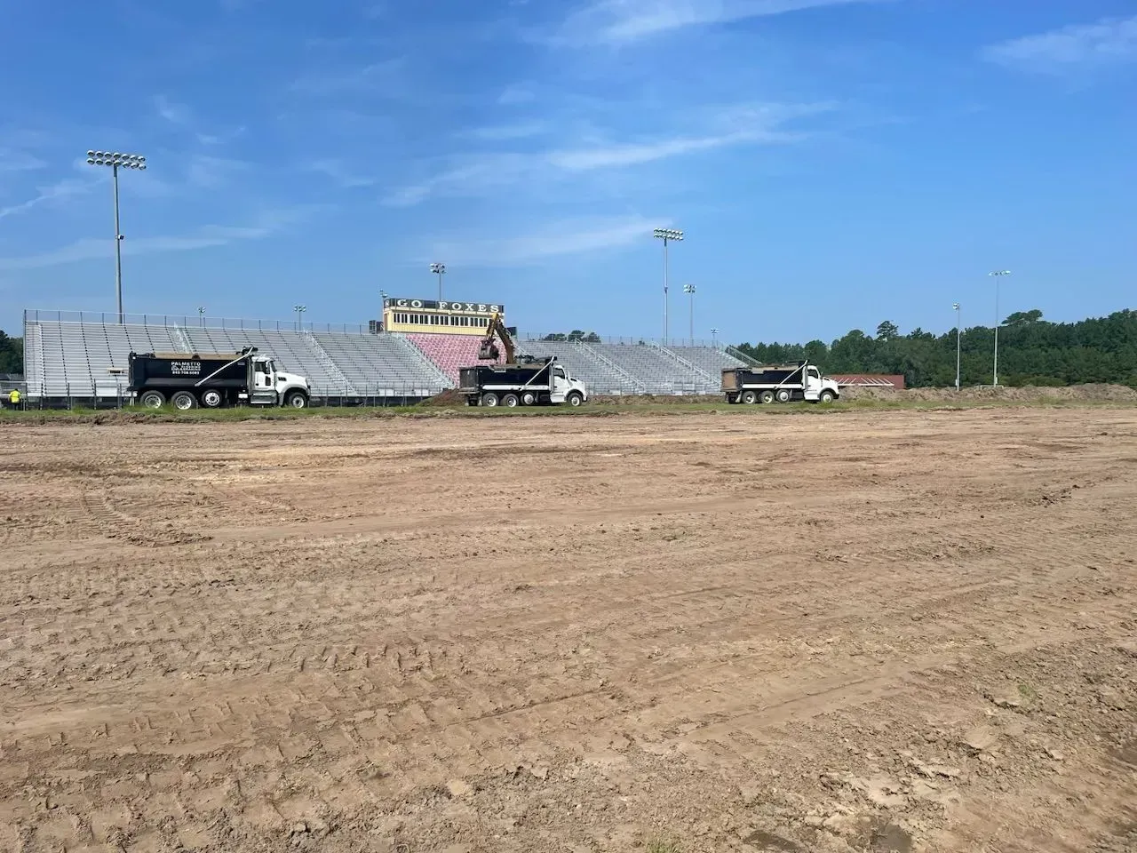 Construction site with three dump trucks in front of a stadium under clear blue sky.