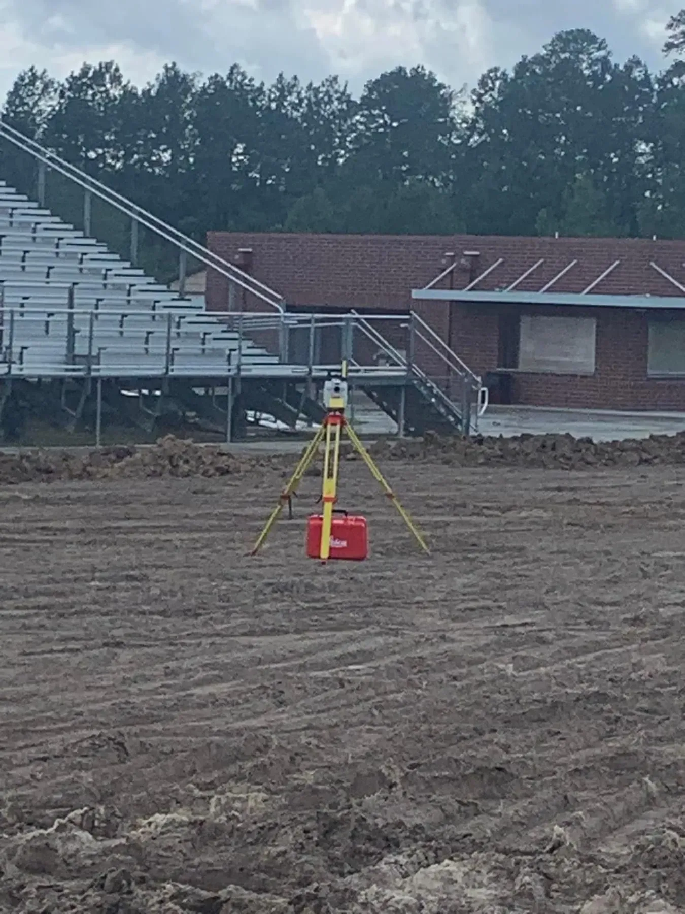 Surveying equipment on a dirt field near stadium seating and a brick building.
