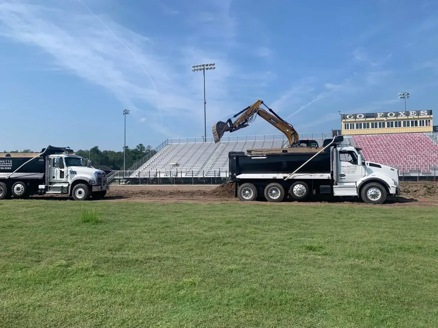 Two dump trucks and excavator near a stadium removing dirt, sunny day.