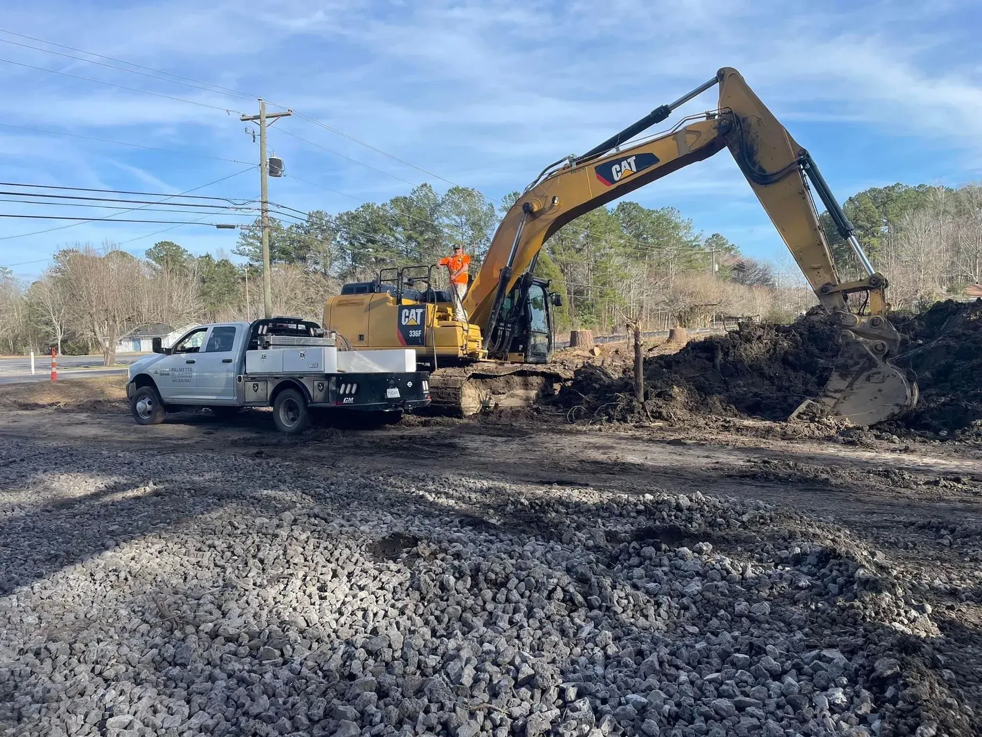 Excavator digging dirt near a white pickup truck on a gravel road, trees in the background, blue sky.