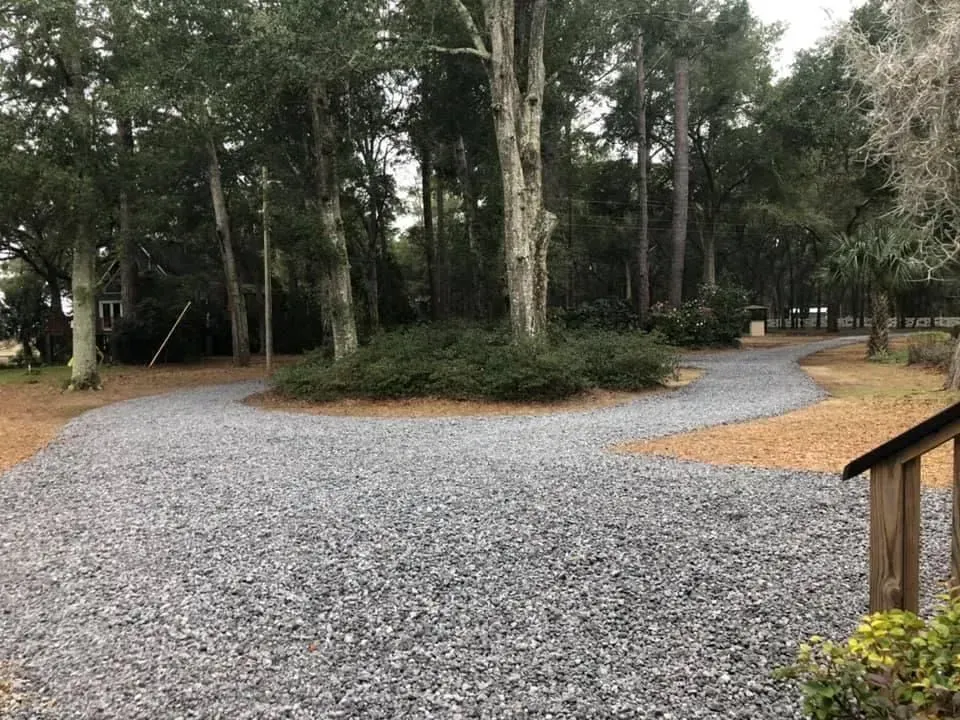 Gravel driveway curves into a shaded, wooded area. Dark gravel, tan dirt, and green bushes.