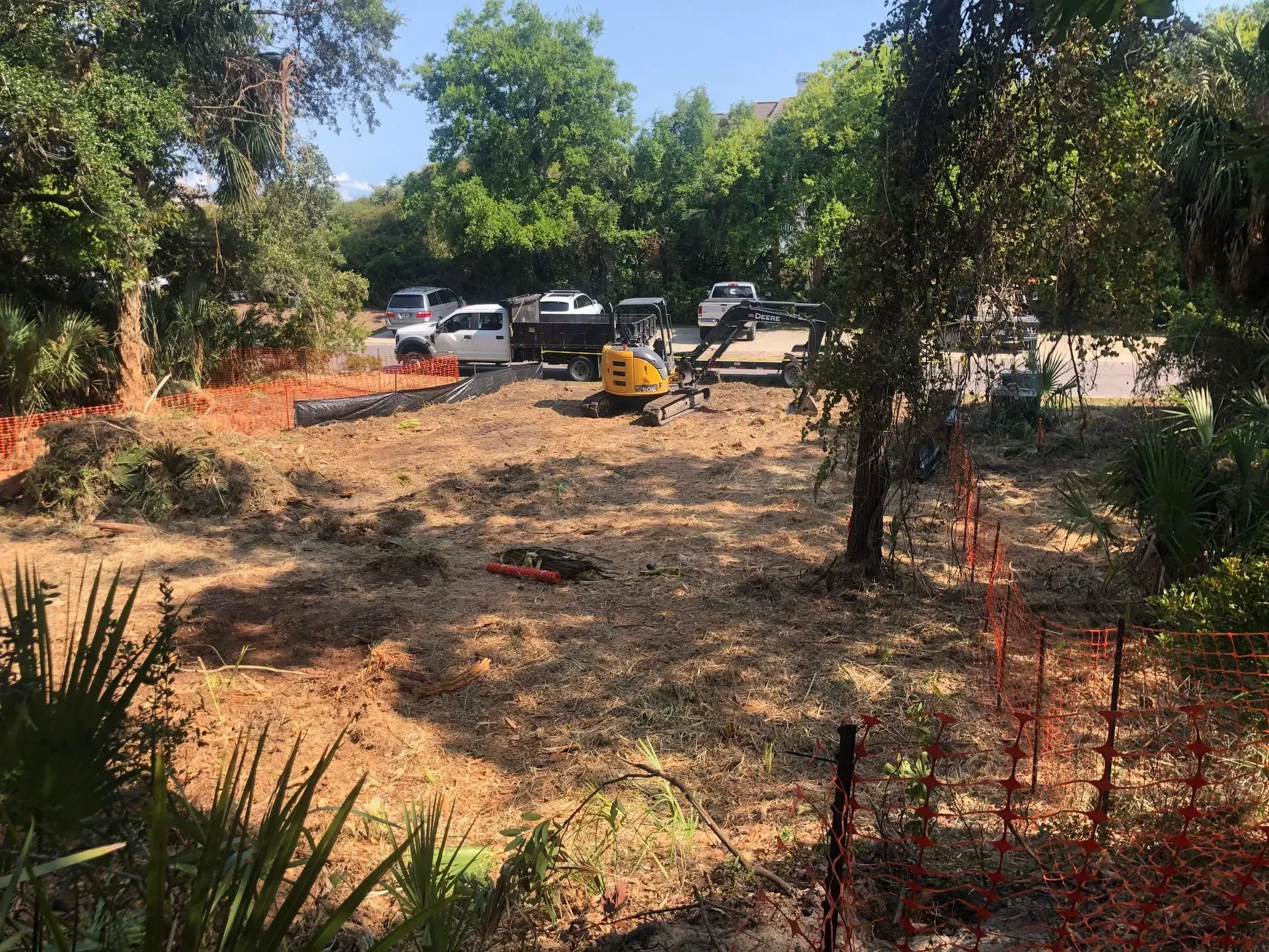 Construction site with excavator and trucks among trees. Orange safety fencing borders the area.