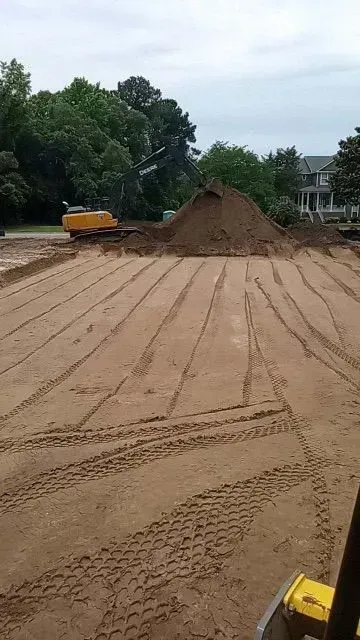 Construction site with dirt piles and an excavator, ground marked with tire tracks and lines.