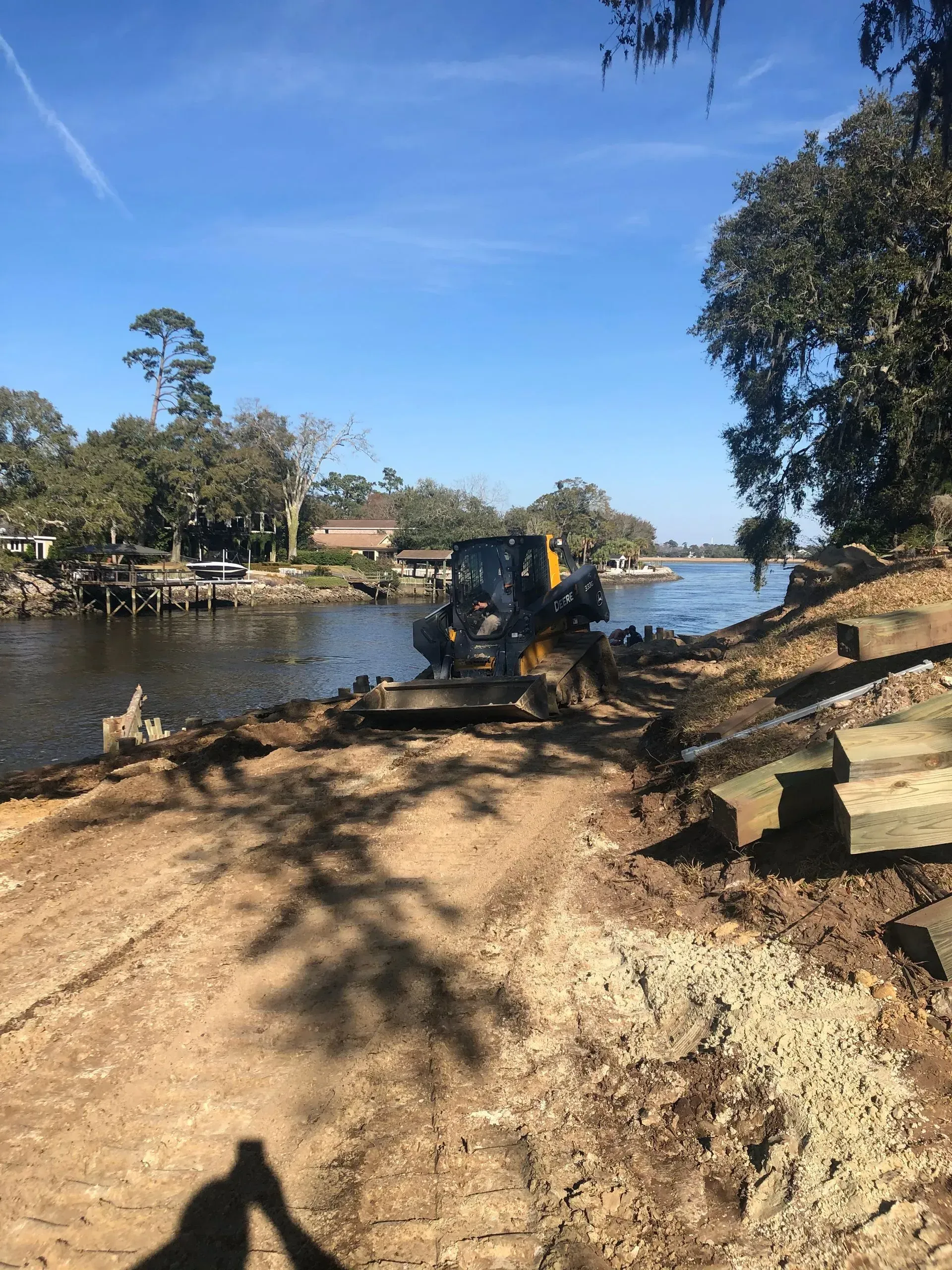 Excavator on a riverbank, removing wooden planks. Clear, sunny day.