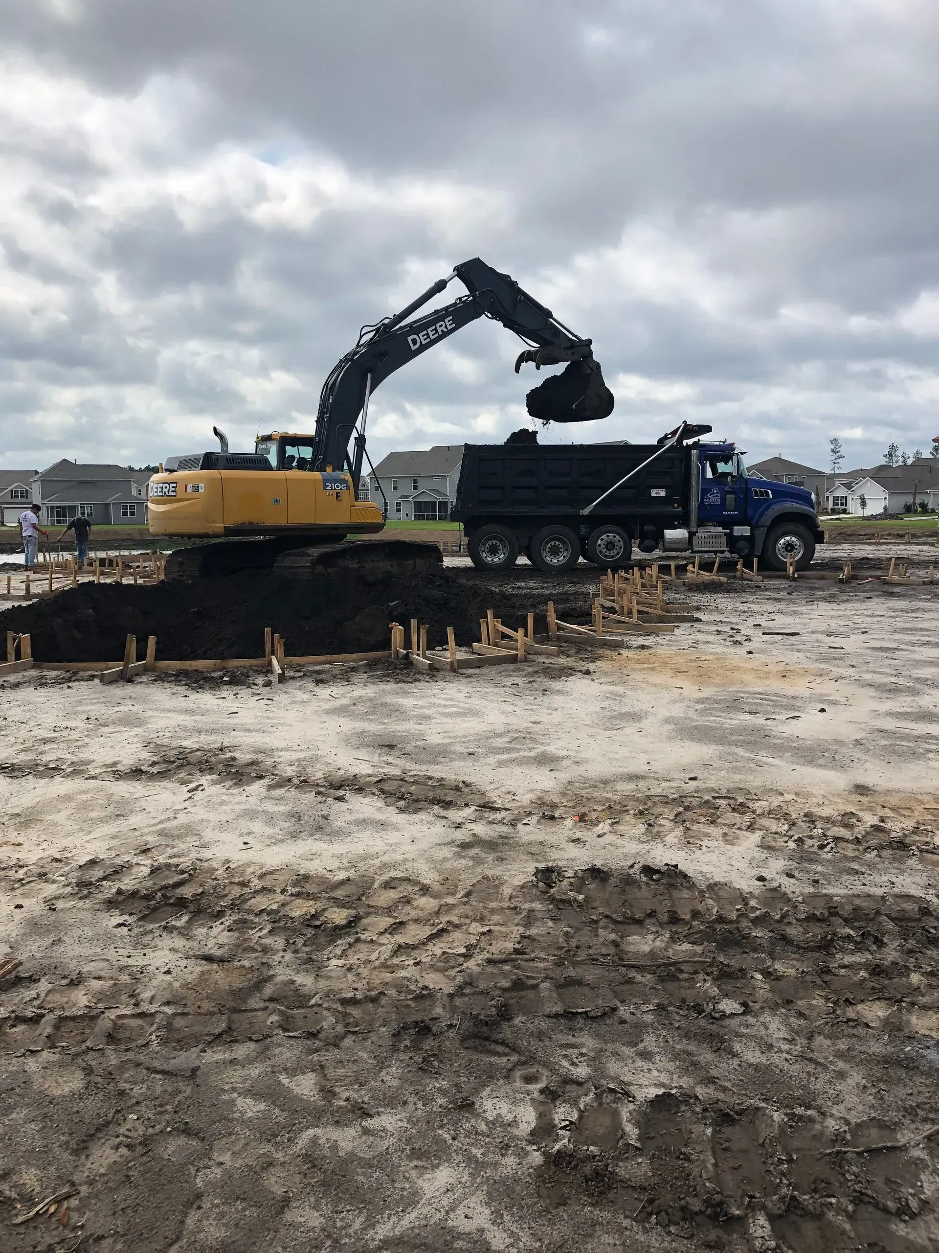 Excavator loading dirt into a dump truck on a construction site under a cloudy sky.