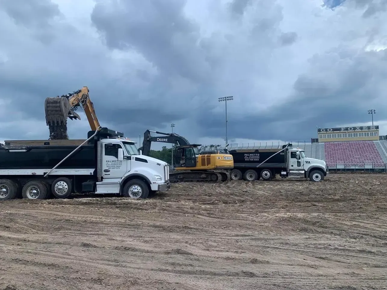 Excavator loading dirt into dump trucks at a construction site near a stadium on an overcast day.