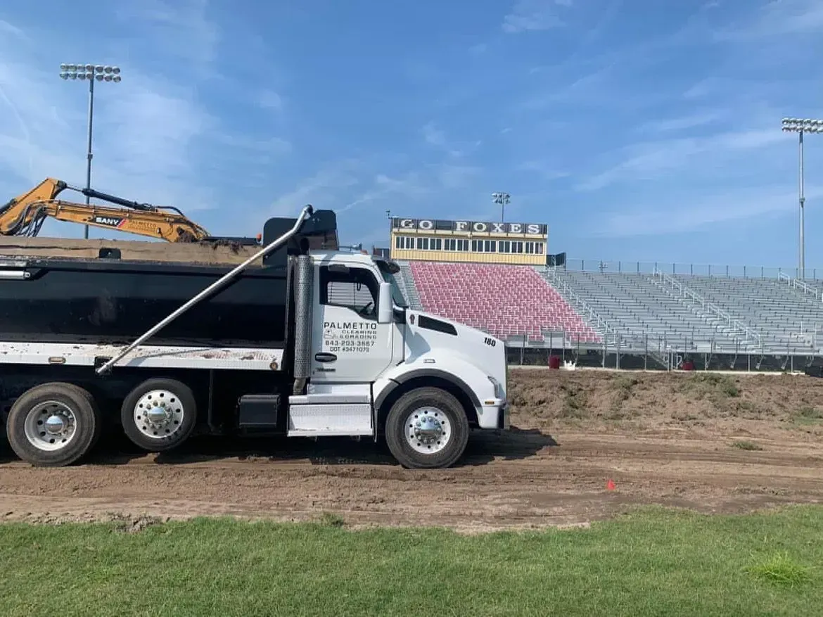 A white dump truck on a dirt field with a stadium in the background. Construction is ongoing.