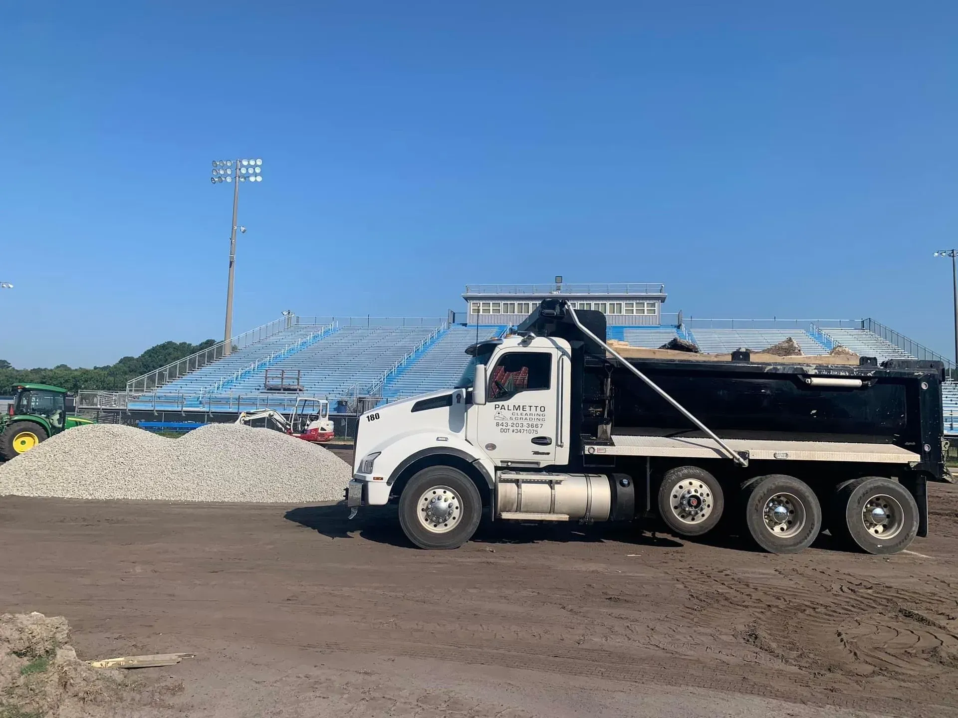 White dump truck hauling rocks near stadium bleachers on a sunny day.