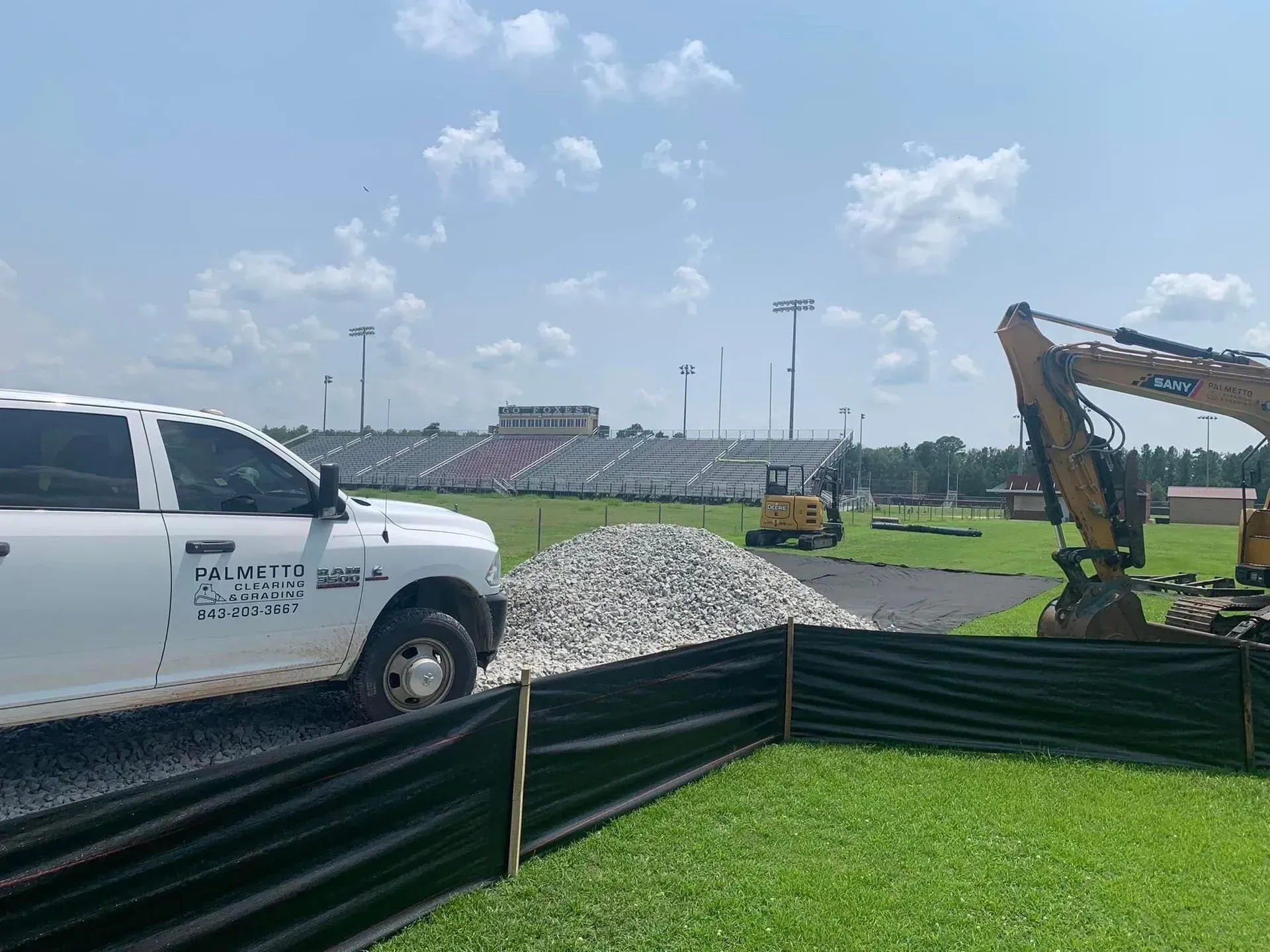 Construction site with a truck, excavator, and stadium in the background under a blue sky.