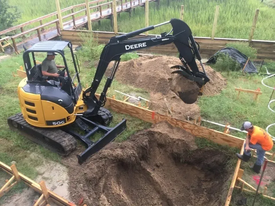 Yellow John Deere excavator digging near a wooden fence, with a worker in orange vest.