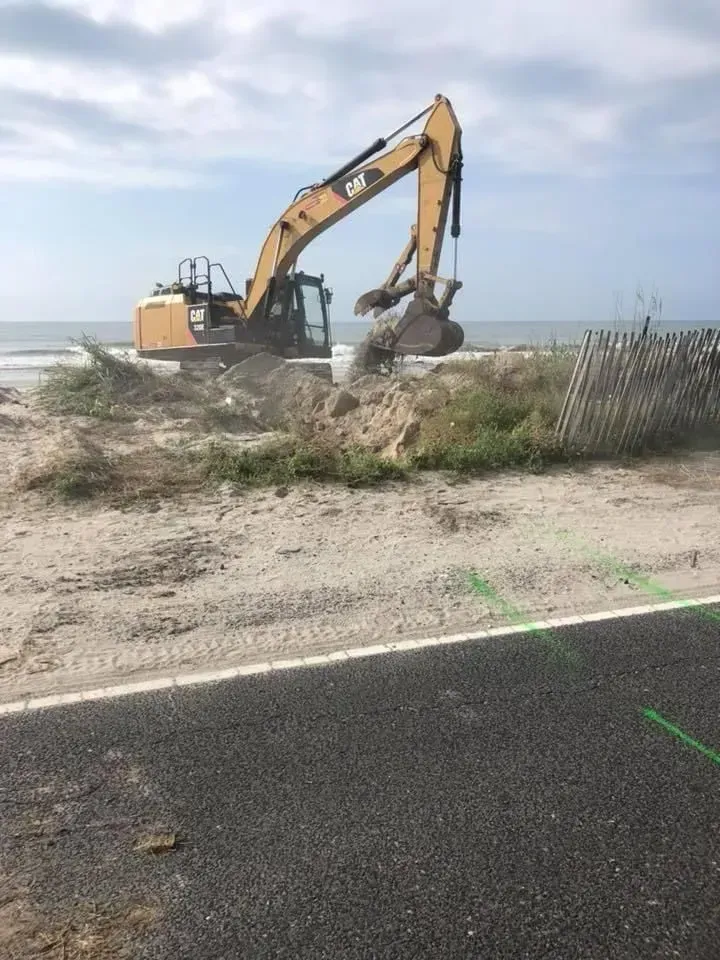 Excavator digging sand near a beach and road. Cloudy sky.