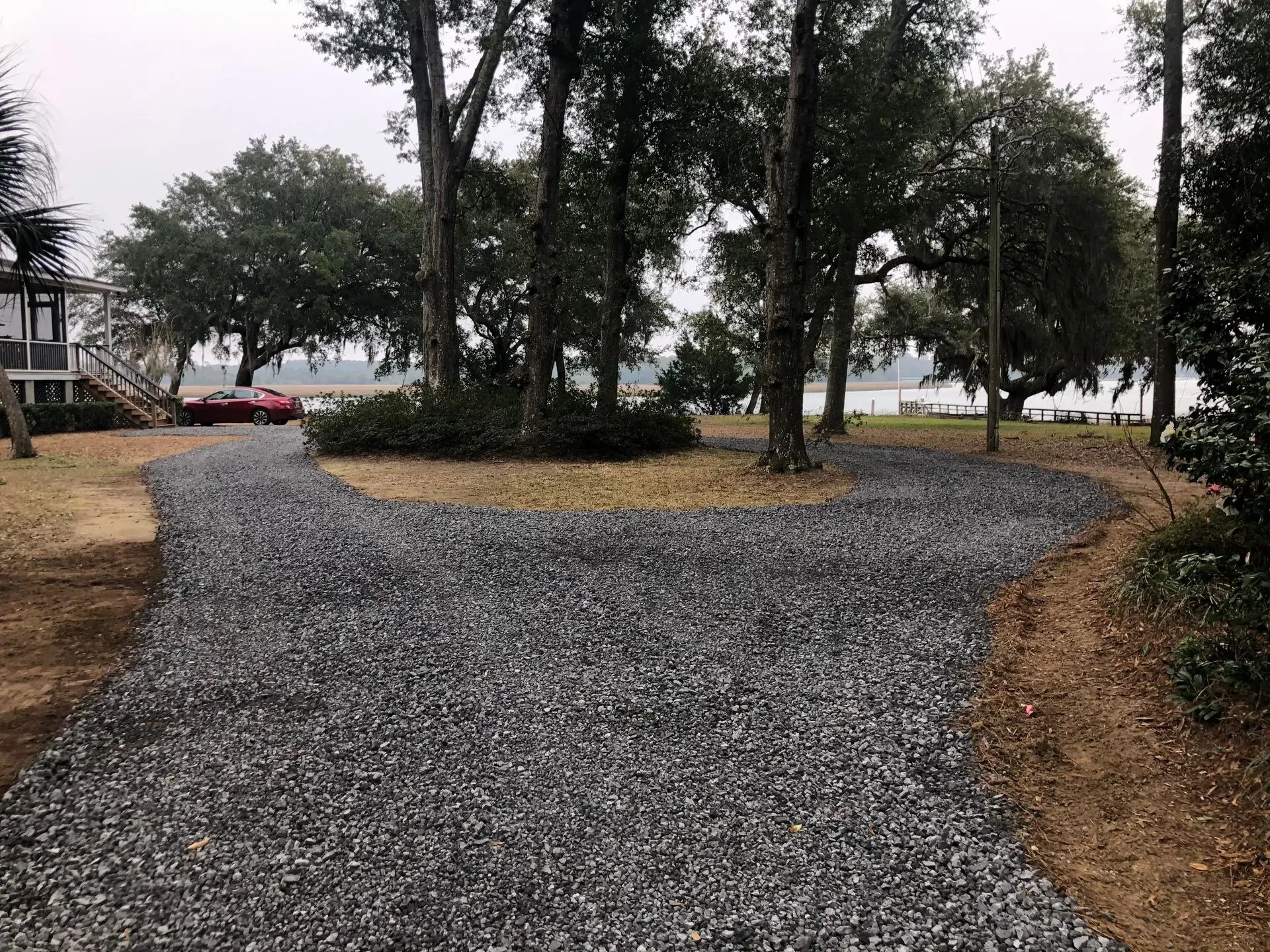 Gravel driveway splitting into two paths around trees, near water, with a red car in the distance.