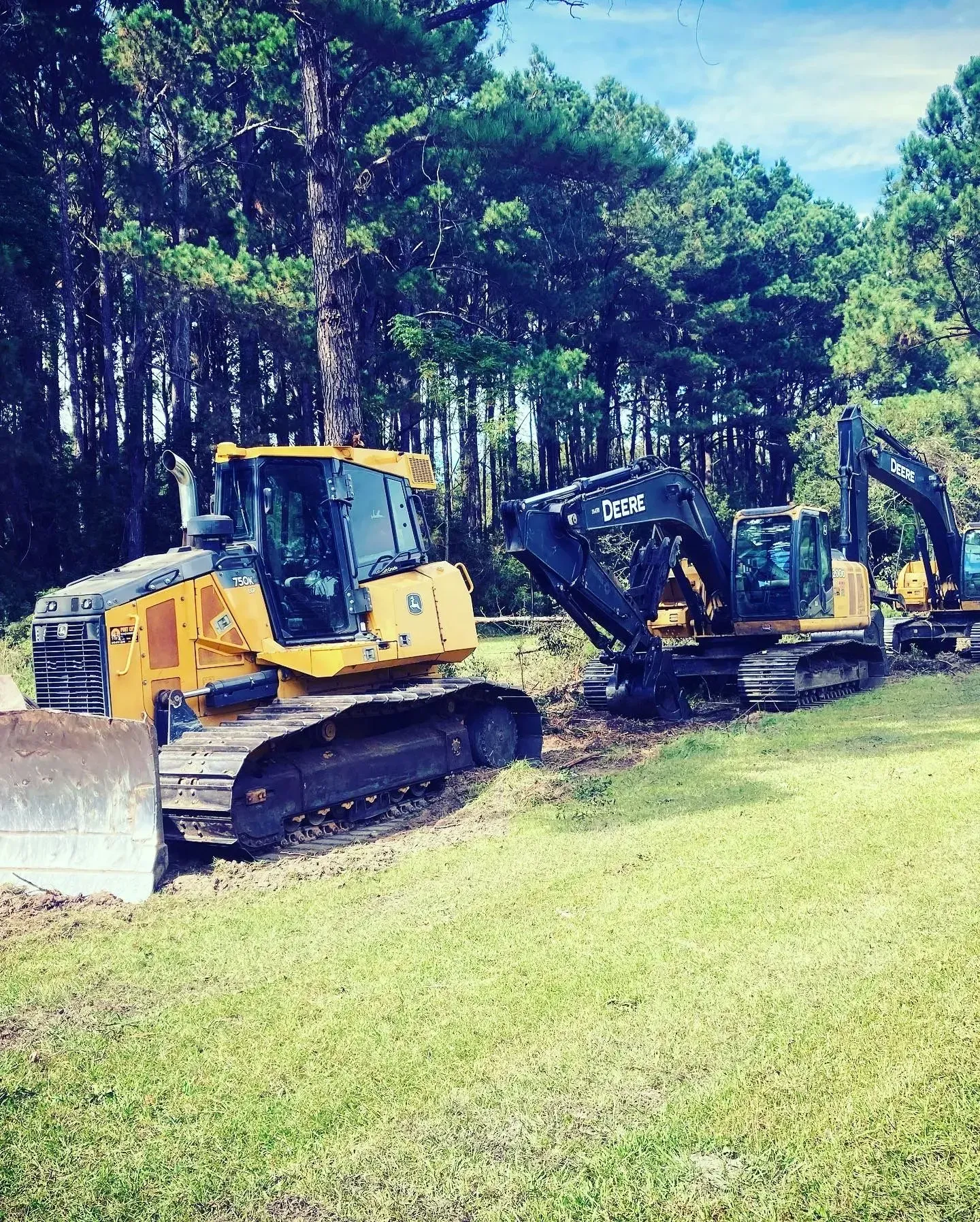 Yellow bulldozer and two excavators parked on grass near a wooded area.