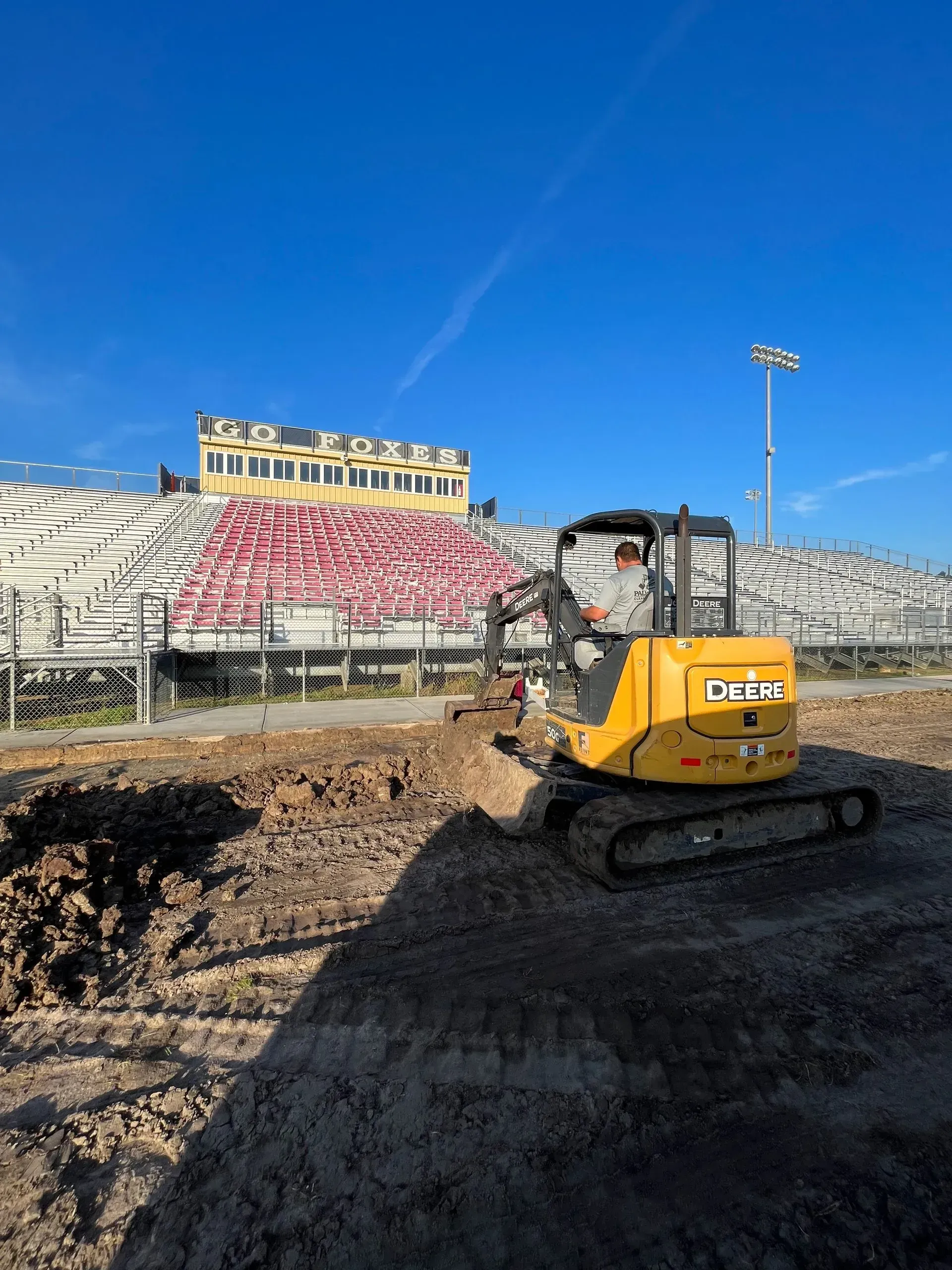 Yellow excavator at a racetrack, digging into the earth. Bleachers and blue sky in the background.