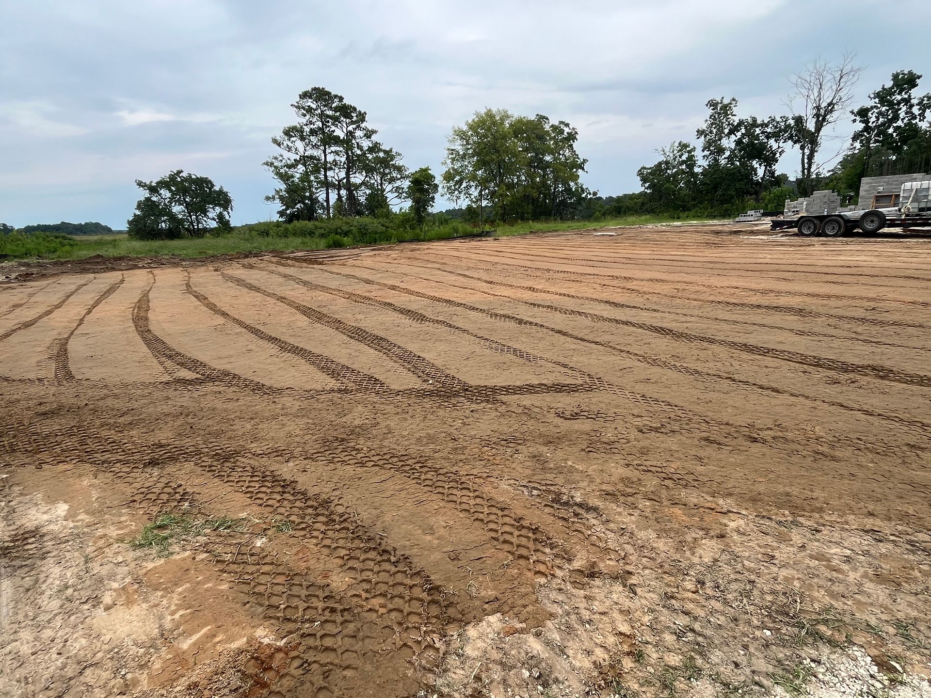 Cleared, dirt-covered construction site with vehicle tire tracks and trees in the background under a cloudy sky.