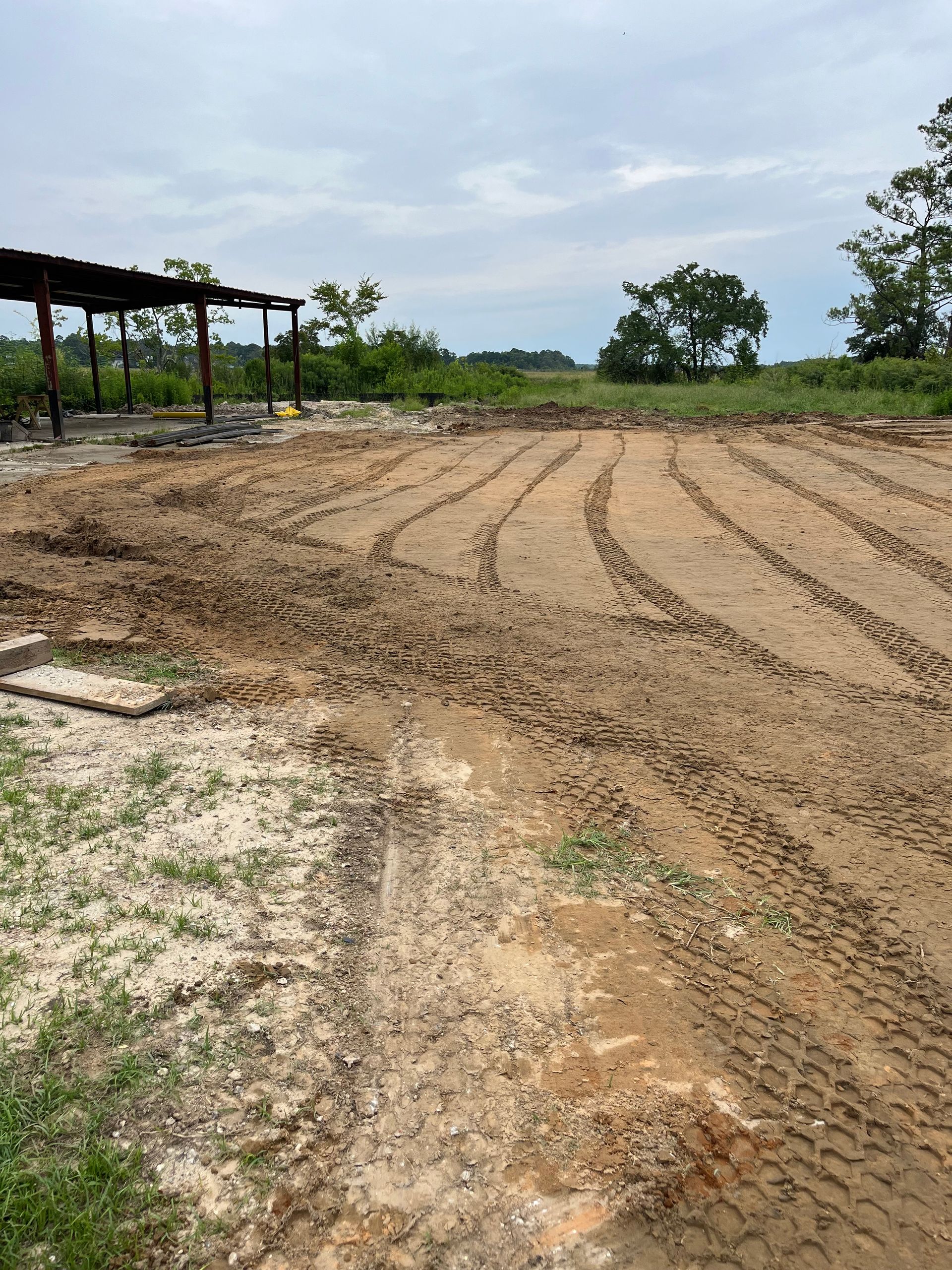 A cleared, muddy construction site with tire tracks, an open-sided metal structure, and distant trees under a cloudy sky.