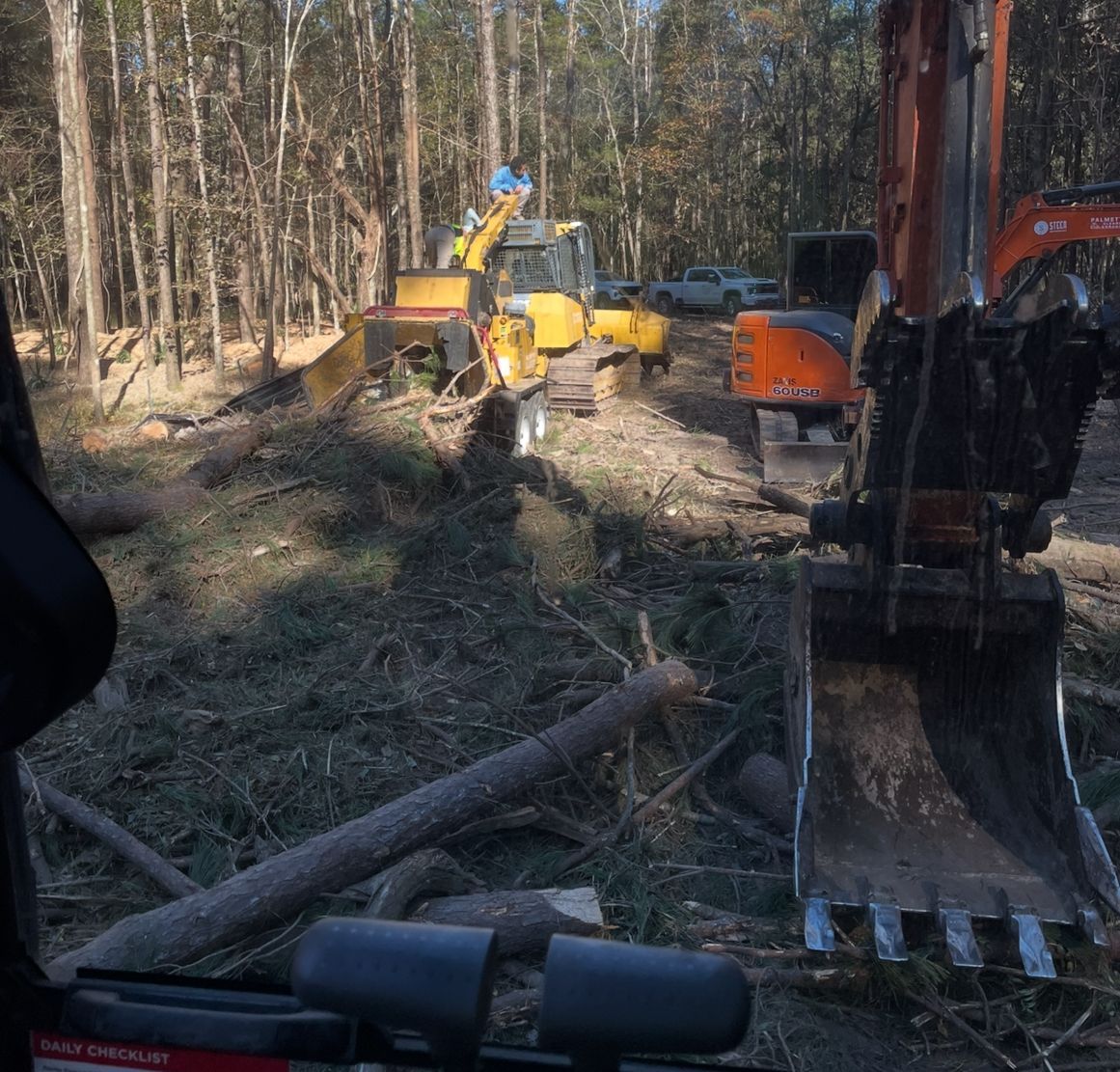 Two excavators work in a wooded area, clearing trees and debris. One yellow machine faces a large orange machine.