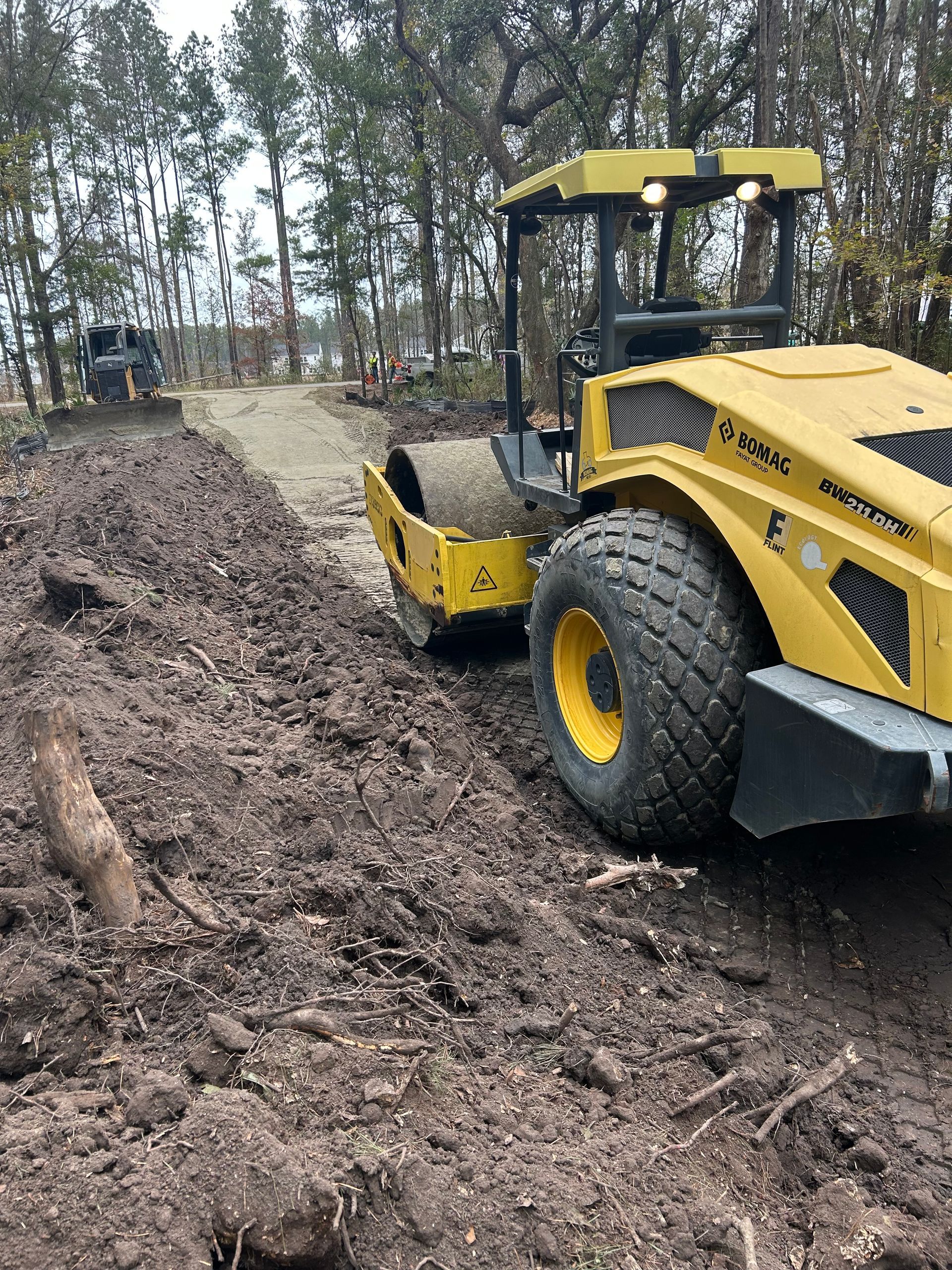 A yellow soil compactor sits on a muddy, unpaved construction path through a wooded area.