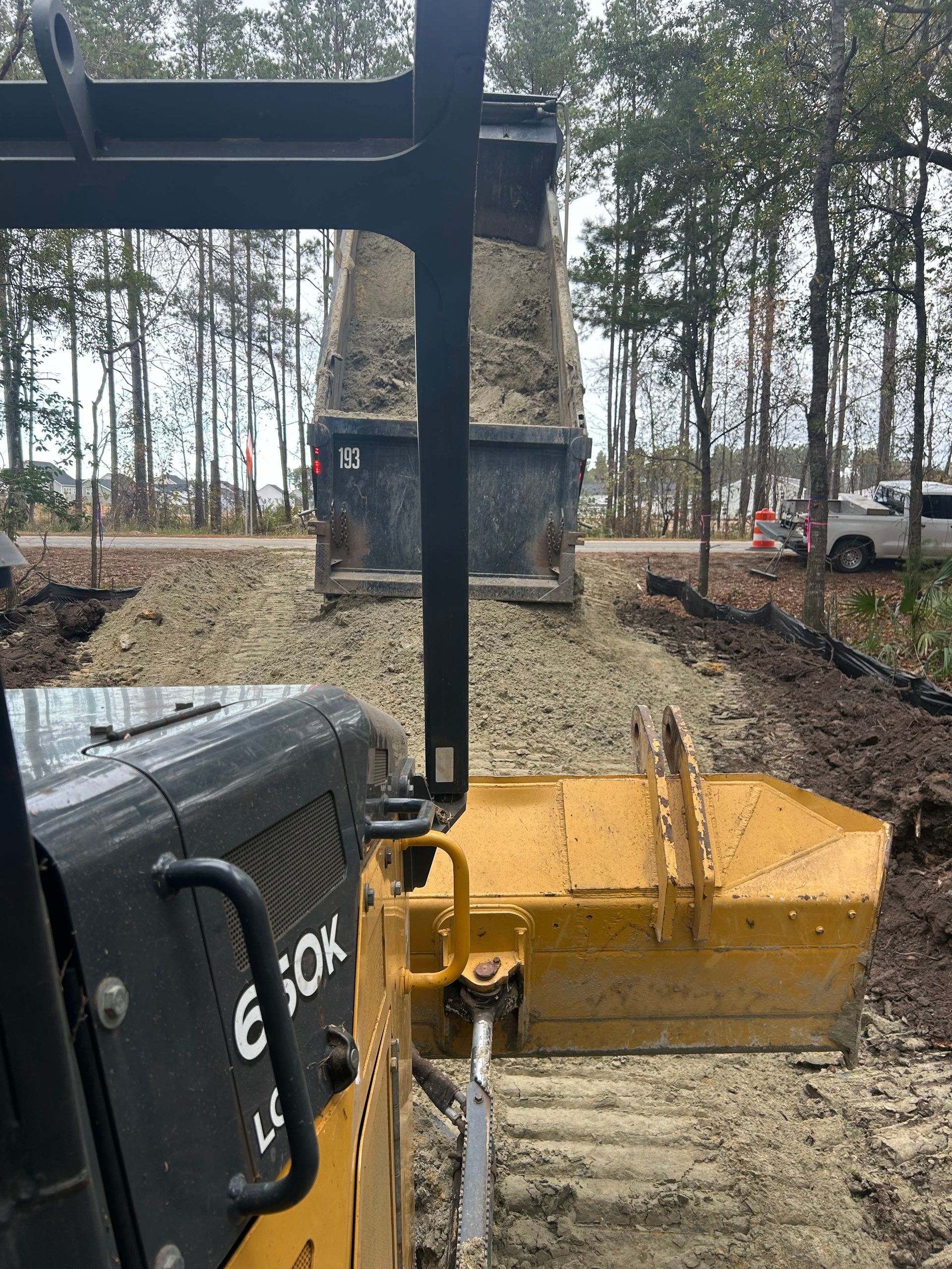 A view from the cab of a yellow bulldozer behind a dump truck spreading gravel on a dirt road in a wooded area.