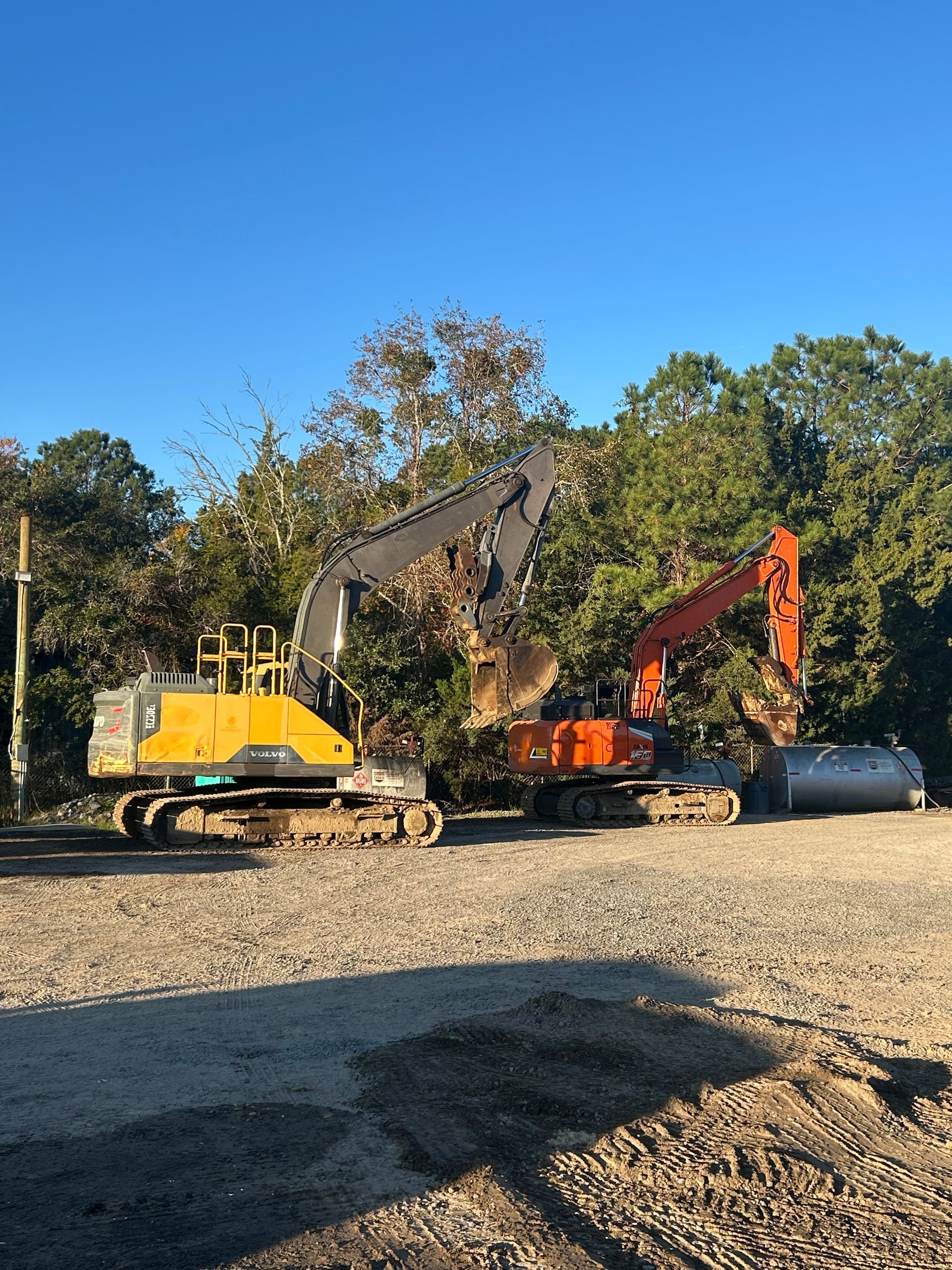 Two excavators, one yellow and one orange, parked on a gravel lot in front of trees under a clear blue sky.